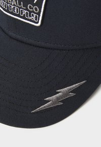 Jūreivio baseball švilpikas su tekstūruota medžiaga, su balta etikete su prekės ženklu ir sidabriniu žaibo siuvinėjimu ant skydelio.