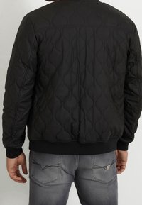 Chaqueta acolchada negra con un patrón texturizado y ondulado, con puños y dobladillo acanalados. Combinada con jeans grises, mostrando detalles traseros y costuras.