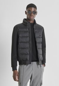 Hombre vestido con una chaqueta acolchada negra con mangas negras, una camiseta negra, pantalones de carga grises y gafas de sol de montura transparente, frente a un fondo gris liso.