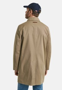 Beige jas met een zachte textuur, voorzien van een klassieke kraag, een split aan de achterkant en leren accent bij de hals. Gedragen over blauwe jeans.