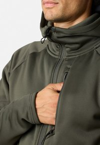 Giacca in pile verde oliva con colletto alto e cappuccio regolabile, dotata di zip frontale e tasca laterale, morbida al tatto e vestibilità aderente.