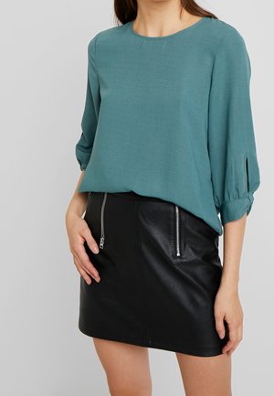 Vrouw draagt een teal driekwart mouw top met splitdetail en een zwarte minirok van nep leer met ritssluitingen aan de voorkant.