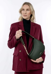 Grüne Lederhandtasche mit geschwungener Form, strukturiertem Oberflächen und einem Goldverschluss. Das Modell hält die Tasche vor einem weißen Hintergrund.