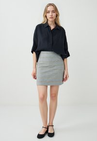 Chemise noire en satin oversized à manches courtes associée à une jupe ajustée en tartan gris et des ballerines noires, sur un fond uni.