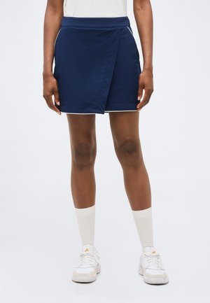 Vrouw die een marineblauwe wikkelrok draagt, witte enkelsokken, witte sneakers en een wit topje, staand tegen een effen achtergrond.