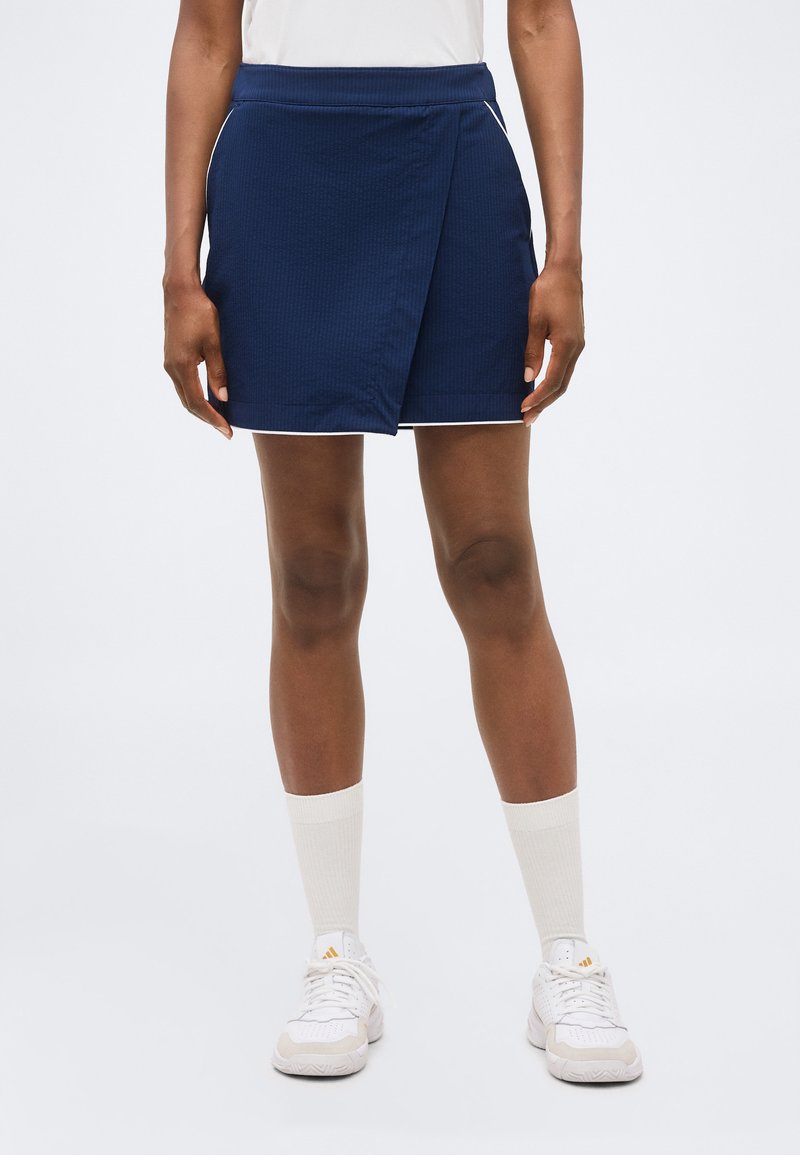 Γυναίκα που φοράει μπλε σκούρη φούστα τύπου wrap, λευκές κάλτσες μέχρι τον αστράγαλο, λευκά αθλητικά παπούτσια και λευκό τοπ, στέκεται μπροστά σε απλό φόντο.