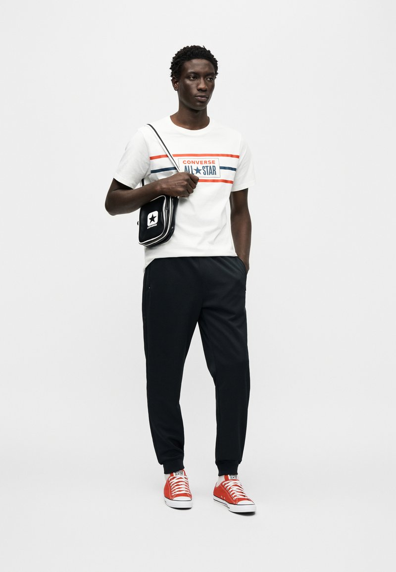 Hvid T-shirt med røde og blå striber og "CONVERSE ALL STAR" logo, sorte bukser og røde sneakers. Lille sort crossbody-taske med logo.