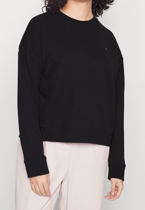 Person trägt ein schlichtes schwarzes Sweatshirt mit langen Ärmeln und helle beige Hosen, steht vor einem einfarbigen Hintergrund.