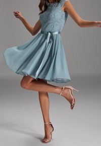 Ein ärmelloses, hellblaues Cocktailkleid mit einem Spitzentop und einem glatten Rock, versehen mit einem gebundenen Taillenbereich und kombiniert mit beigen Riemchen-Heels.