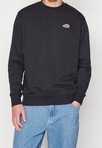 Svart bomulls sweatshirt med rund hals, långa ärmar, och en liten röd och vit sneakers-logotyp på vänster bröst. Tillsammans med blåjeans.