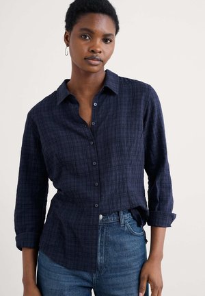 Donna con capelli corti che indossa una camicia blu navy con bottoni e maniche arrotolate, insieme a jeans blu, in piedi di fronte a uno sfondo chiaro e semplice.