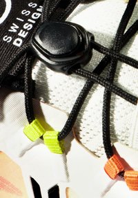 Scarpa bianca in rete con lacci neri e un toggle nero. Caratterizzata da accenti colorati: clips di fissaggio verdi e arancioni. Il testo dice "DESIGN SVIZZERO."