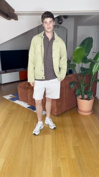 Gaiši zaļš žakets virs brūnas krekla, balti šorti un pelēkas kedas. Iekštelpu vide ar puķu podu un dīvānu fona.