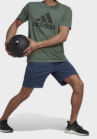 Homme en t-shirt Adidas vert et shorts bleus tenant un ballon médicinal Adidas noir, les jambes écartées, portant des baskets Adidas noires.