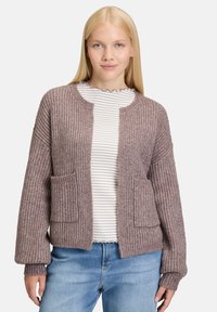 Młoda kobieta z prostymi blond włosami, ubrana w ribbed fioletową kardigan nad białą bluzką w paski i niebieskimi dżinsami, stojąca na jednolitym tle.