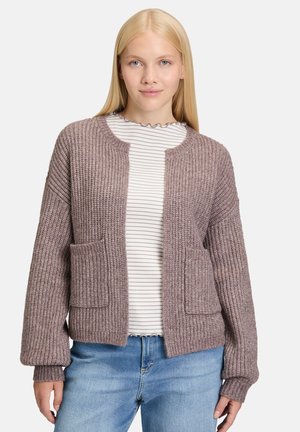 Jeune femme aux cheveux blonds raides portant un cardigan mauve à côtes sur un haut rayé blanc et un jean bleu, se tenant devant un fond uni.