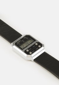 Montre numérique avec un bracelet en cuir noir et un boîtier rectangulaire en argent. L'écran présente un fond noir avec des chiffres et des boutons blancs.