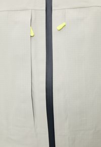 Tela gris con una cremallera negra central y dos bolsillos verticales, cada uno con una pequeña pestaña de tirador amarilla parcialmente visible.