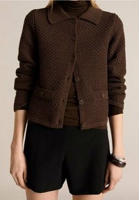 Bruine gebreide cardigan met een kraag, knoopsluiting en zakken. Draag je over een donkere top en gecombineerd met zwarte shorts.