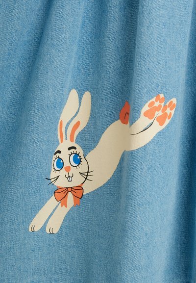 Lapin blanc de dessin animé aux yeux bleus, longues oreilles et nœud papillon orange, pattes étirées, sur fond de tissu bleu clair.