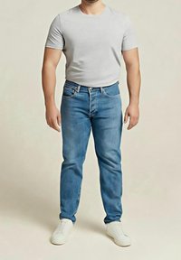 Homme debout portant un t-shirt gris clair à manches courtes, un jean bleu et des baskets blanches sur un fond uni.