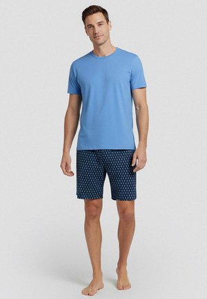 Uomo in piedi scalzo che indossa una camicia azzurro chiaro a maniche corte e pantaloncini blu navy con fantasia su uno sfondo semplice.