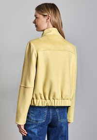 Veste en daim synthétique jaune avec un col montant, une taille élastiquée et des manches longues. Conçue pour une coupe décontractée.