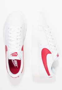 Vita Nike-sneakers med ett texturerat läderövre, med en framträdande röd Swoosh-logotyp, vita snören och en röd inredning.