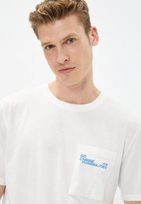 Camiseta de algodón blanca con cuello redondo y mangas cortas. Presenta un bolsillo en el lado izquierdo del pecho con texto azul: "VVE A BUSCAR SUS POSIBILIDADES".