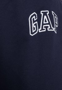 Navyblå bomullssweatshirt med vit broderad "GAP"-logotyp i blockbokstäver. Enkelt och strukturerat tyg. Närbild.
