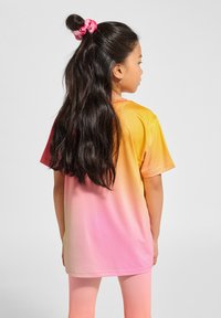 Kortärmad T-shirt i en gradient från gult till rosa, tillverkad av ett smidigt tyg. Håret är stylat med en rosa scrunchie. Inga synliga mönster.