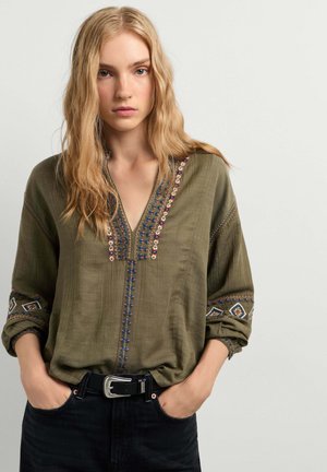 Joven con cabello rubio largo, vestida con una blusa verde oliva bordada y vaqueros negros, con las manos en los bolsillos, sobre un fondo liso.