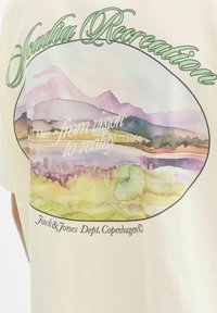 Krēms krāsas kokvilnas T-krekls ar akvareļu ainavu grafiku, kurā attēloti kalni un upe, papildināts ar zaļu tekstu un melnām kontūrām.