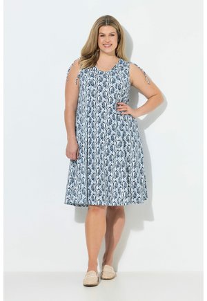 Smilende kvinde i ærmeløs knælængde blå og hvid mønstret kjole med bindestropper, stående med hånd på hoften iført beige slip-on sko.