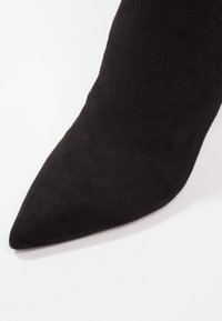 Stivaletto nero in suede con punta affusolata, texture liscia, silhouette affilata e tacco leggermente curvato, concentrandosi su un design moderno ed elegante.