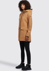 Giacca impermeabile marrone con cappuccio, chiusura frontale con zip, due grandi tasche e dettagli neri. Indossata con jeans skinny neri e sneakers.