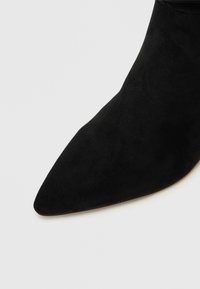 Bottes noires en daim à cheville avec un talon pointu et une texture lisse. Le design présente une silhouette élégante et une esthétique minimaliste.