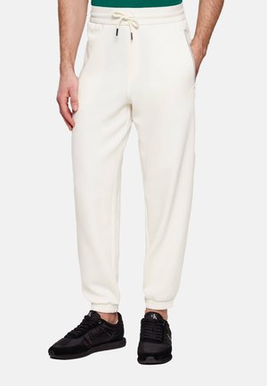 Uomo che indossa pantaloni da jogging bianchi con polsini elastici e vita con coulisse, abbinati a scarpe da ginnastica nere e una maglietta verde.