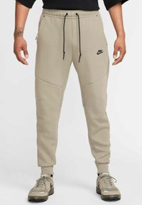Beige Nike Jogginghosen mit elastischem Bund und schwarzen Kordeln, ausgestattet mit schrägen Seitentaschen und dezenten Nähten.