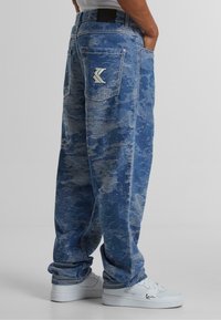 Blue denim pants featuring a camo pattern, relaxed fit, and visible stitching. Includes two back pockets and a logo detail on the waistband.