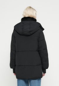 Person med kort blondt hår iført en svart polstret frakk med hette og fuskepelskrage, kombinert med blå jeans, vist bakfra.