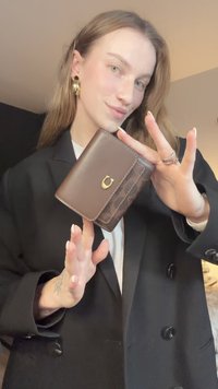Portefeuille en cuir marron avec un accent en métal doré ; présente une surface texturée et un design compact. Le modèle le tient avec un blazer noir.