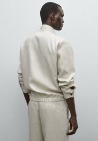 Hellbeige Jacke mit Stehkragen, hochgekrempelten Ärmeln und elastischem Bund. Glatte Textur, minimalistisches Design, kombiniert mit passenden Hosen.