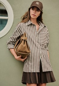 Camicia a righe con bottoni marrone e crema, abbinata a una gonna marrone. La modella tiene una borsa in camoscio color cognac con dettaglio fibbia.