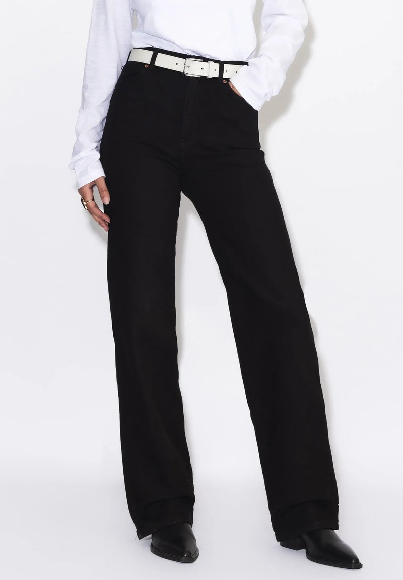 Persona con pantalones negros de tiro alto y pierna ancha, cinturón blanco, camisa blanca de manga larga, zapatos negros puntiagudos y mano en el bolsillo.