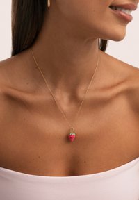 Woman wearing a thin gold chain necklace with a red and green strawberry-shaped pendant, wearing a white off-shoulder top.