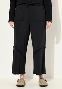 Pantaloni neri a gamba larga realizzati in tessuto morbido, con tasche frontali, orli sfrangiati e pannelli testurizzati a contrasto lungo le gambe.