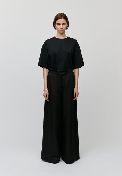 Černé oversized tričko s krátkými rukávy spárované s širokými černými kalhotami, které mají knoflíkové zapínání a hladkou, matnou texturu.