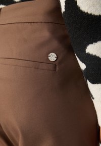 Gros plan sur la poche d'un pantalon marron avec une petite épingle-logo ronde argentée et une partie de la manche d'un pull à motifs noir et blanc.