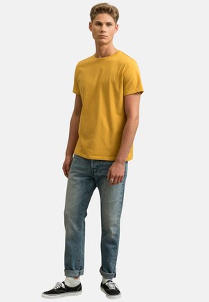Gele katoen T-shirt met korte mouwen, gecombineerd met lichtblauwe denim spijkerbroek. Model draagt zwarte schoenen en witte sokken, staand tegen een effen achtergrond.